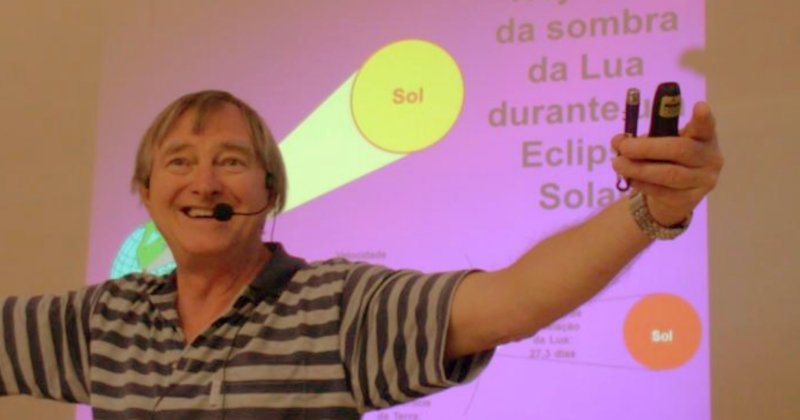 Homem branco de meia idade com microfone de cabeça e os braços abertos em frente à uma tela projetada com tema de astronomia