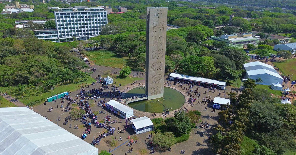 Visão aérea de uma praça com árvores, tendas grandes, uma torre ao centro e várias pessoas circulando