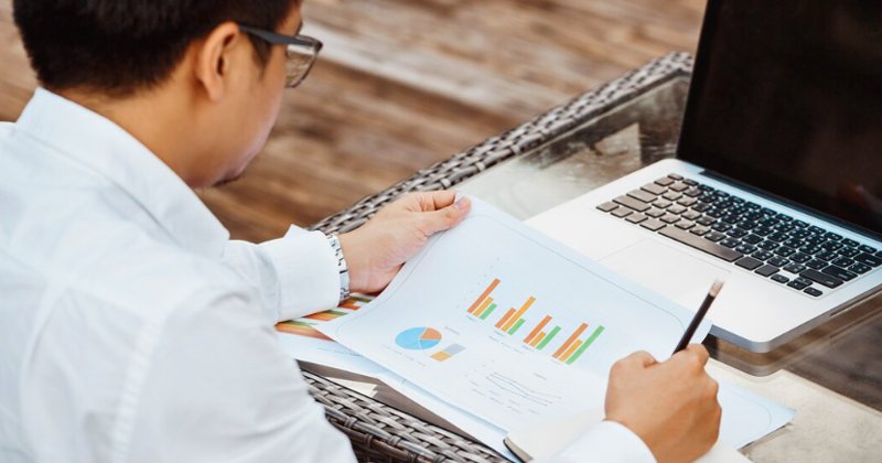 Homem branco de costas com jaleco branco olhando e segurando uma folha com um gráfico e à frente um notebook