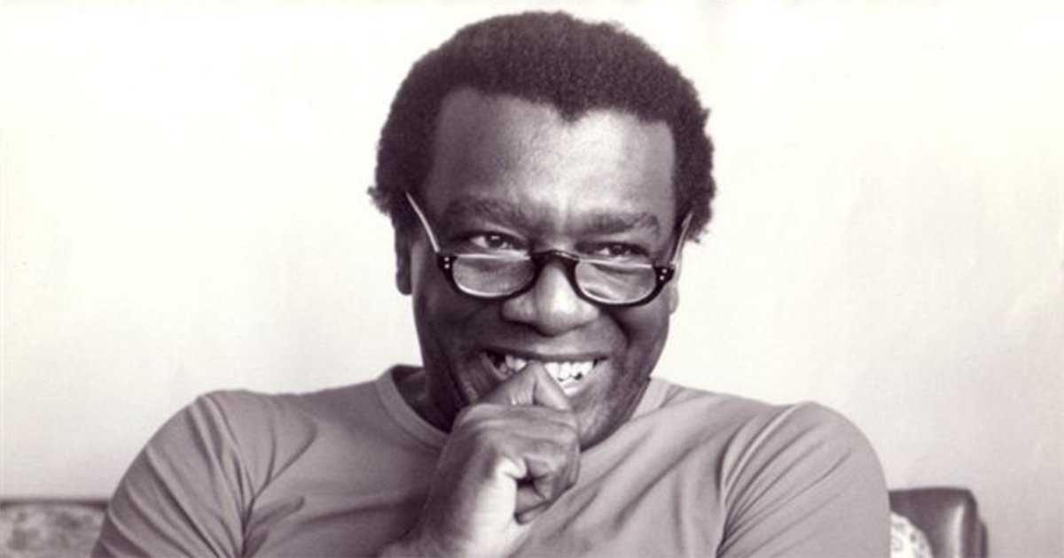 Retrato de Milton Santos sorrindo, com a mão fechada parcialmente em frente à boca. Homem negro, com cabelo curto, óculos de grau e camiseta. Parece estar sentado em um sofá.