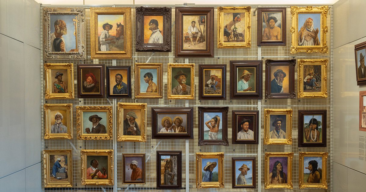 Parede com quadros em molduras marrons e douradas, mostram retratos de pessoas negras