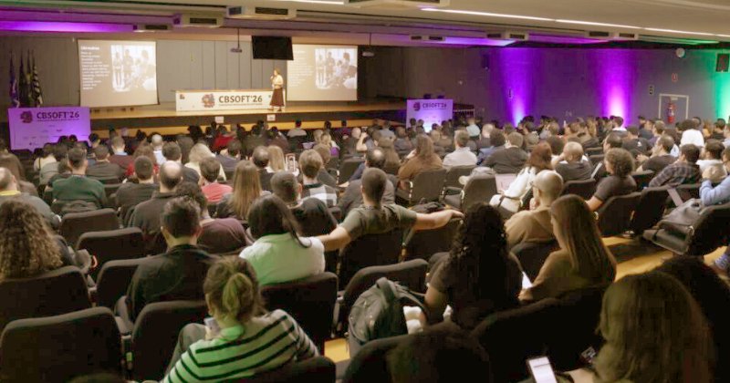 Pessoas sentadas num auditório ouvindo uma palestrante falar no palco ao fundo
