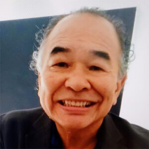 Homem idoso, de cabelos grisalhos e curtos, aparece sorrindo em um retrato. Ele veste uma camisa azul-escura sob um blazer preto e está sentado diante de uma parede de fundo escuro.