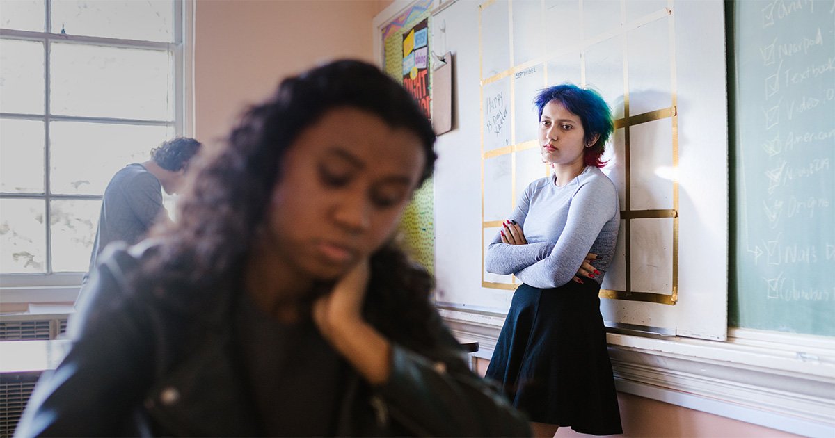 Sala de aula com três estudantes. Em primeiro plano, uma adolescente aparece desfocada, sentada à carteira e apoiando o rosto em uma das mãos. Ao fundo, outra adolescente está encostada em um quadro branco, com os braços cruzados, enquanto um terceiro estudante aparece próximo à janela.