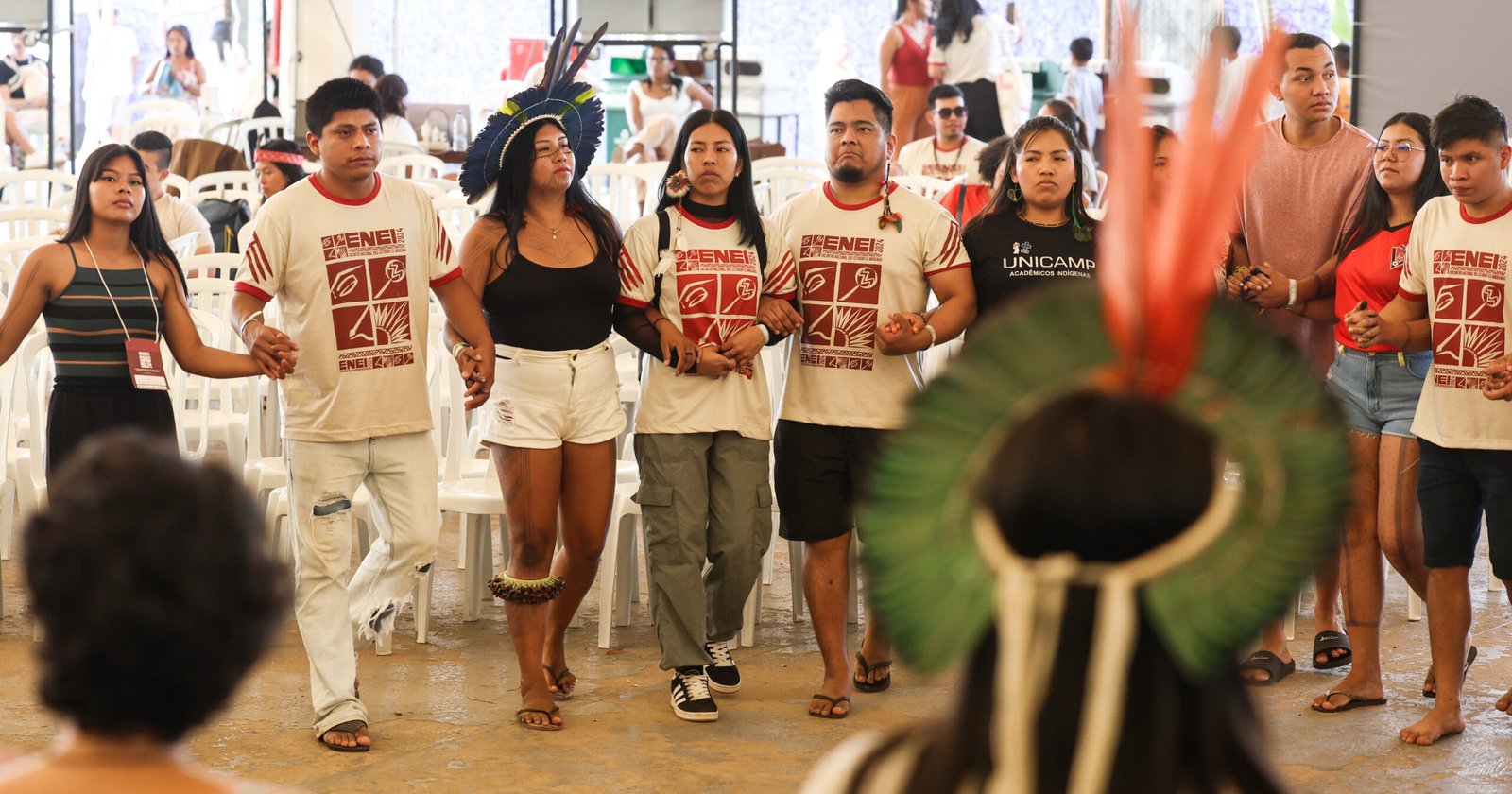 Estudantes indígenas de braços dados em uma roda, em frente a fileiras de cadeiras de plástico. Dois participantes da roda usam cocares de penas. Ao fundo, algumas pessoas conversam em grupos menores.