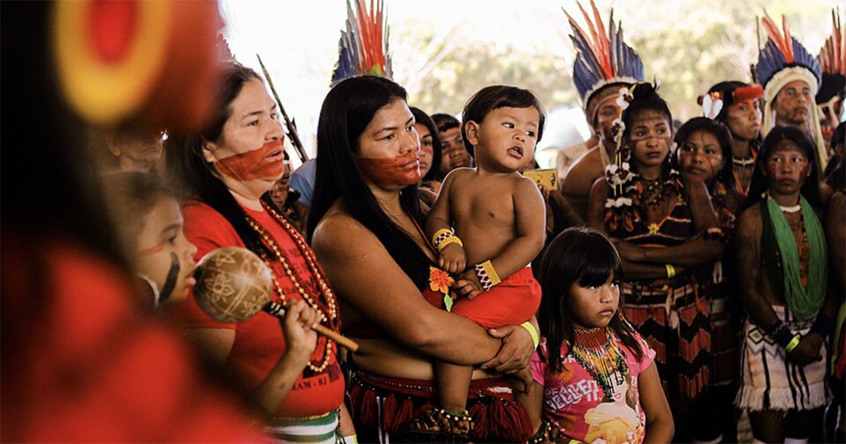 Grupo de pessoas indígenas reunidas em um ambiente aberto. No centro, uma mulher de cabelos longos e pretos segura uma criança pequena no colo, enquanto uma menina está à sua frente. Ao redor, outras pessoas indígenas aparecem usando pinturas corporais, colares, roupas e adornos tradicionais.