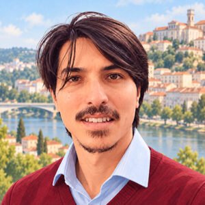 Fotografia de um homem jovem, de cabelos castanhos escuros, lisos e médios, bigode e barba rala no queixo. Ele sorri para a câmera e veste uma camisa azul-clara sob um suéter vinho com decote em V.