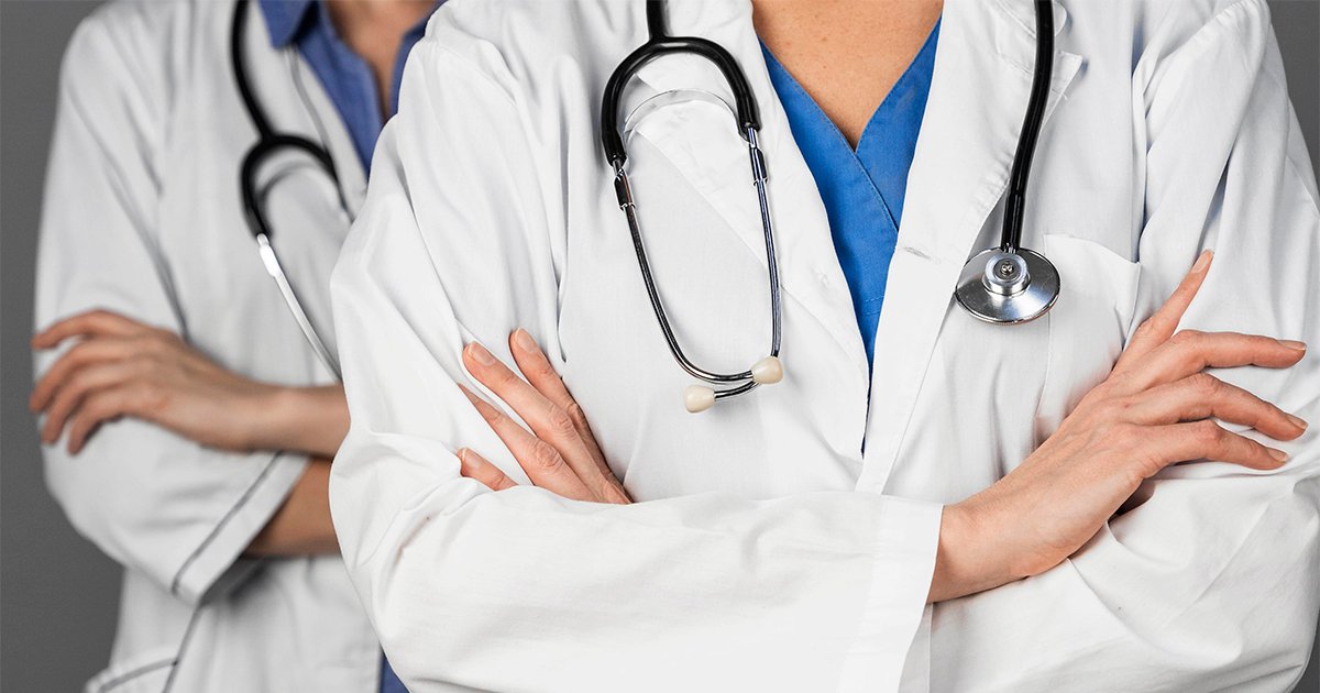 Imagem mostra dois médicos de braços cruzados, com seus estetoscópios, enquadrados do pescoço até a altura do estômago, ou seja, apenas parte do tronco