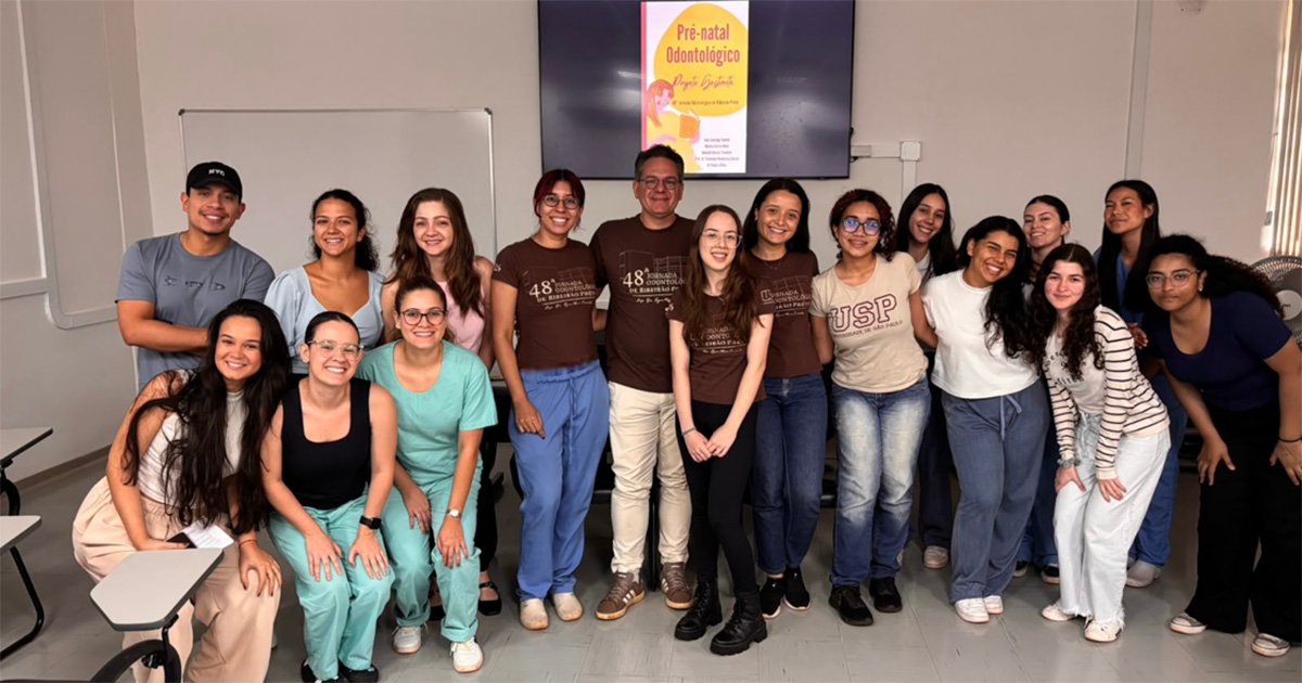 Fotografia num ambiente de sala de aula com um grupo de 16 pessoas sorridentes posicionadas lado a lado. Ao fundo, uma televisão exibe a apresentação do projeto