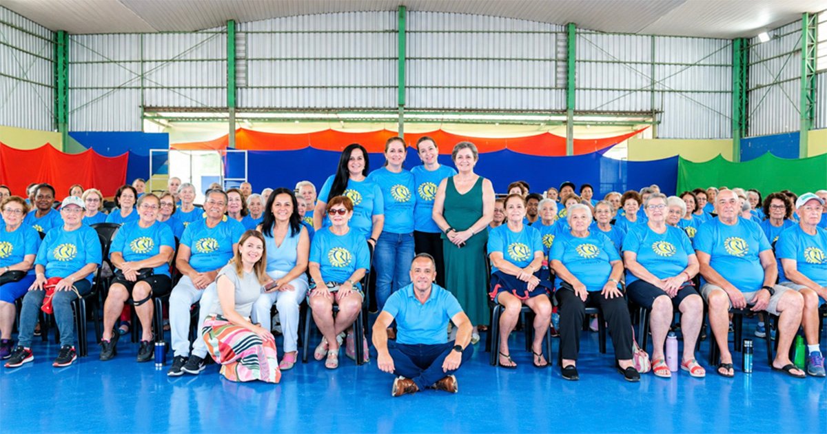 A fotografia mostra um grande grupo de pessoas, predominantemente idosas, reunido dentro de um ginásio ou quadra coberta com piso azul brilhante e teto metálico inclinado.