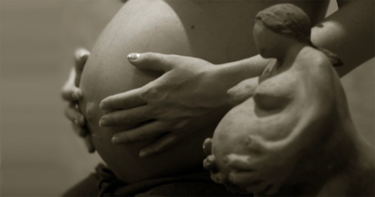 Escultura em tons de sépia representando uma mulher grávida, com as mãos sobre a barriga, enquanto uma segunda pessoa toca seu ventre