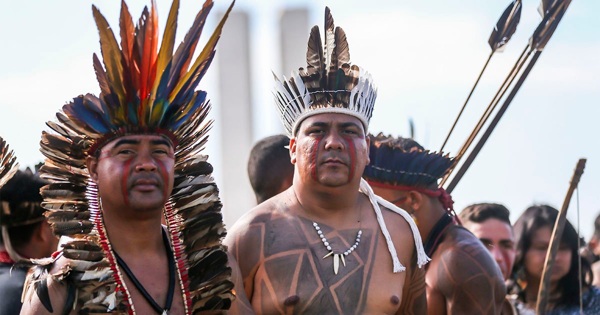 Dois homens indígenas com seus trajes típicos em meio a outros fotografados diante de um prédio em Brasilia - DF