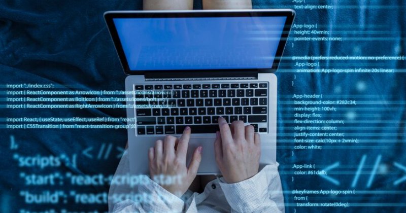 Pessoa digitando em um notebook, visto de cima, com códigos de programação sobrepostos à imagem em tons de azul