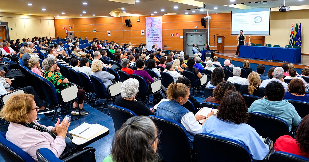 Uma plateia, vista de costas. As cadeiras estão ocupadas. A frente, um palco copm uma mesa azul, um telão branco. As paredes são marrons.